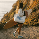 Person carrying a surfboard and a tote bag with 'The Body Not Big Pharma' text on a coastal path.