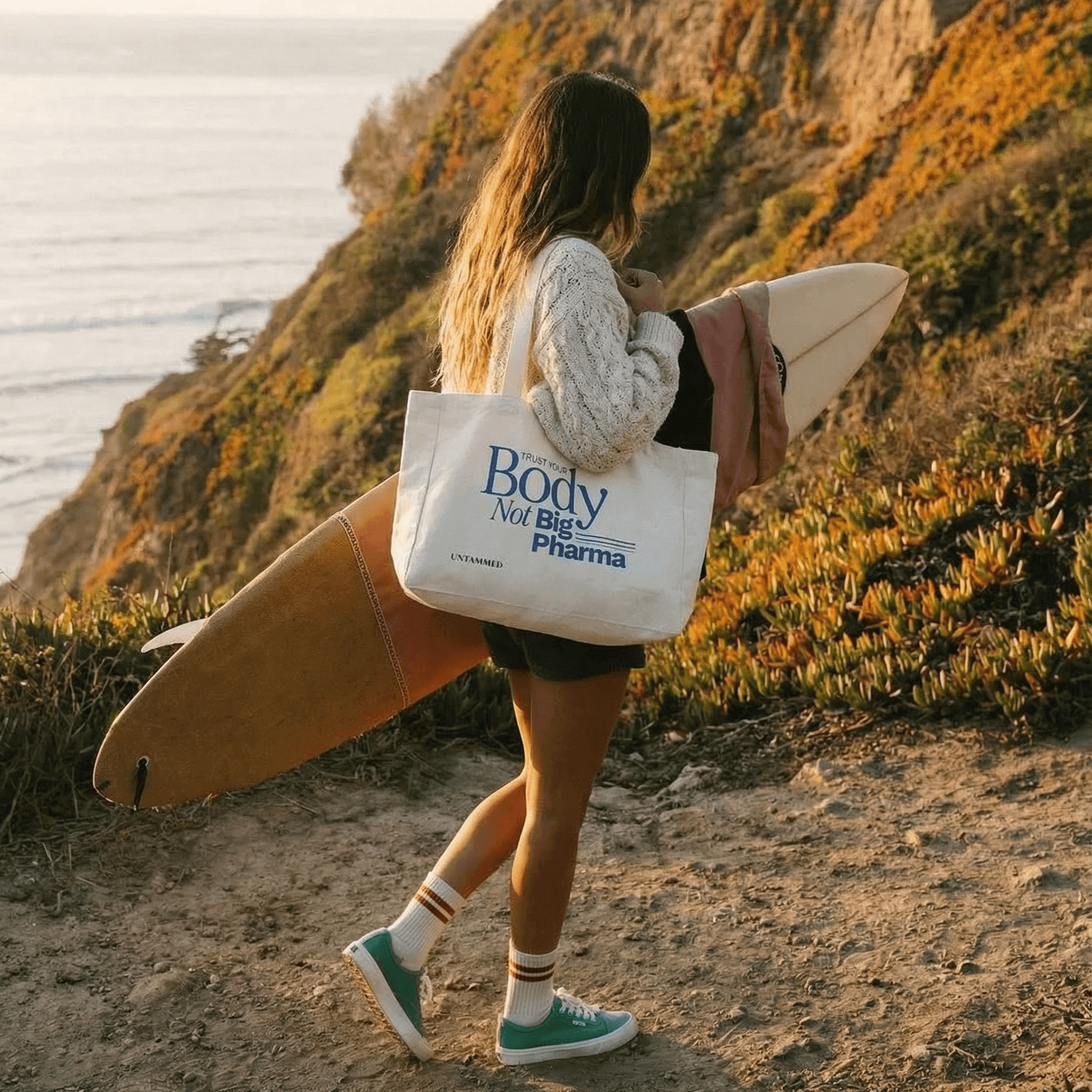 Person carrying a surfboard and a tote bag with 'The Body Not Big Pharma' text on a coastal path.