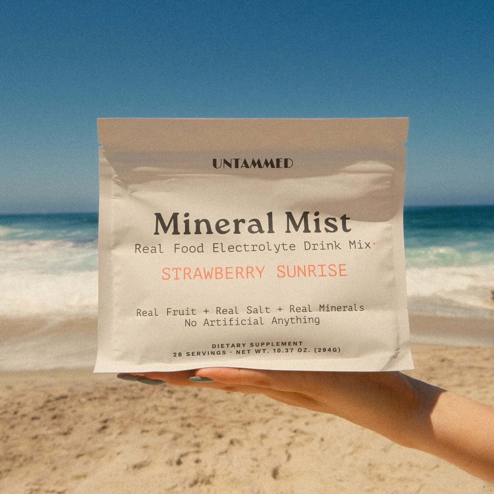 A hand holds a stand-up pouch of UNTAMMED Mineral Mist Strawberry Sunrise electrolyte drink mix against a sunny beach background with sand and ocean waves.