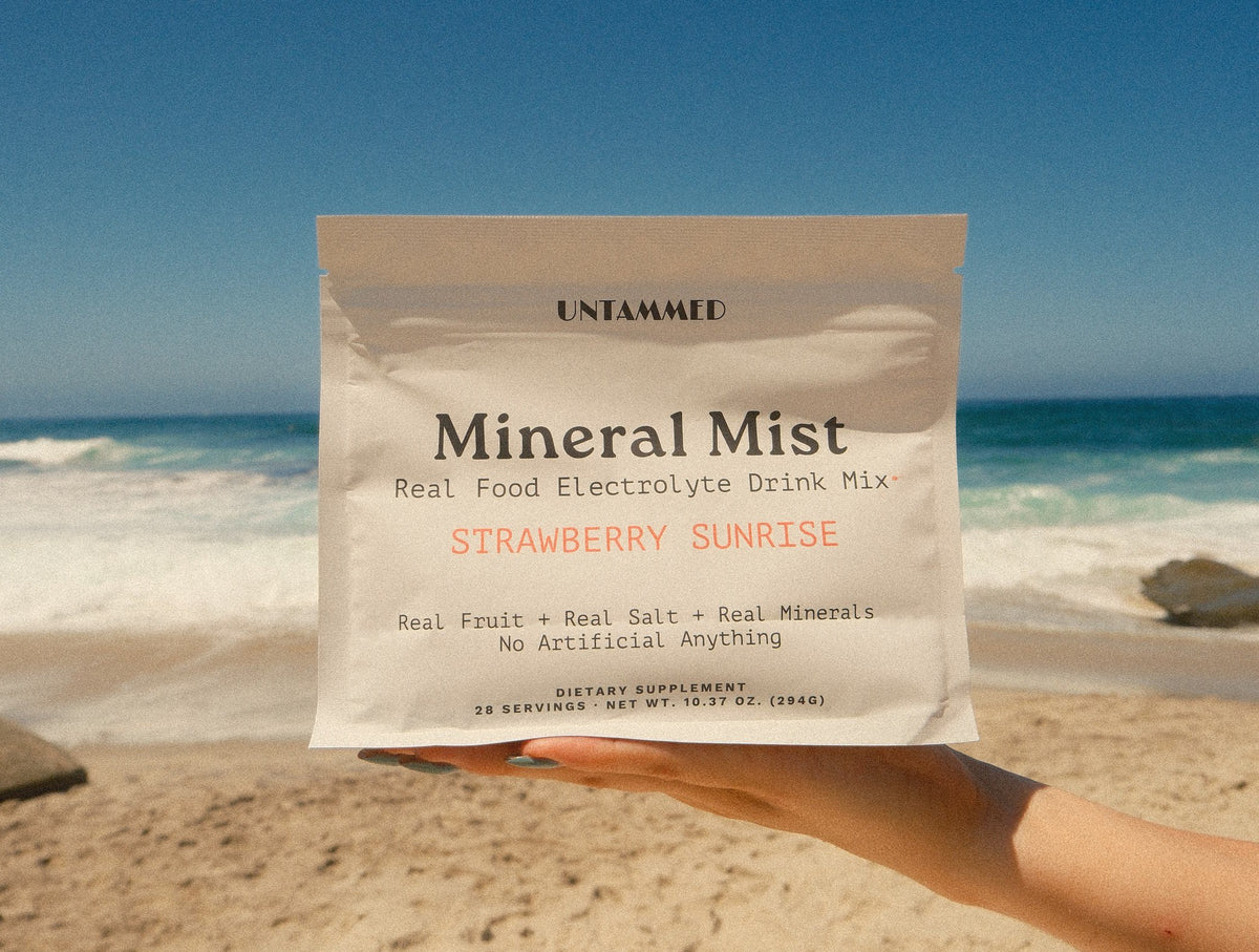 A hand holding a bag of Mineral Mist Strawberry Sunrise up against the ocean